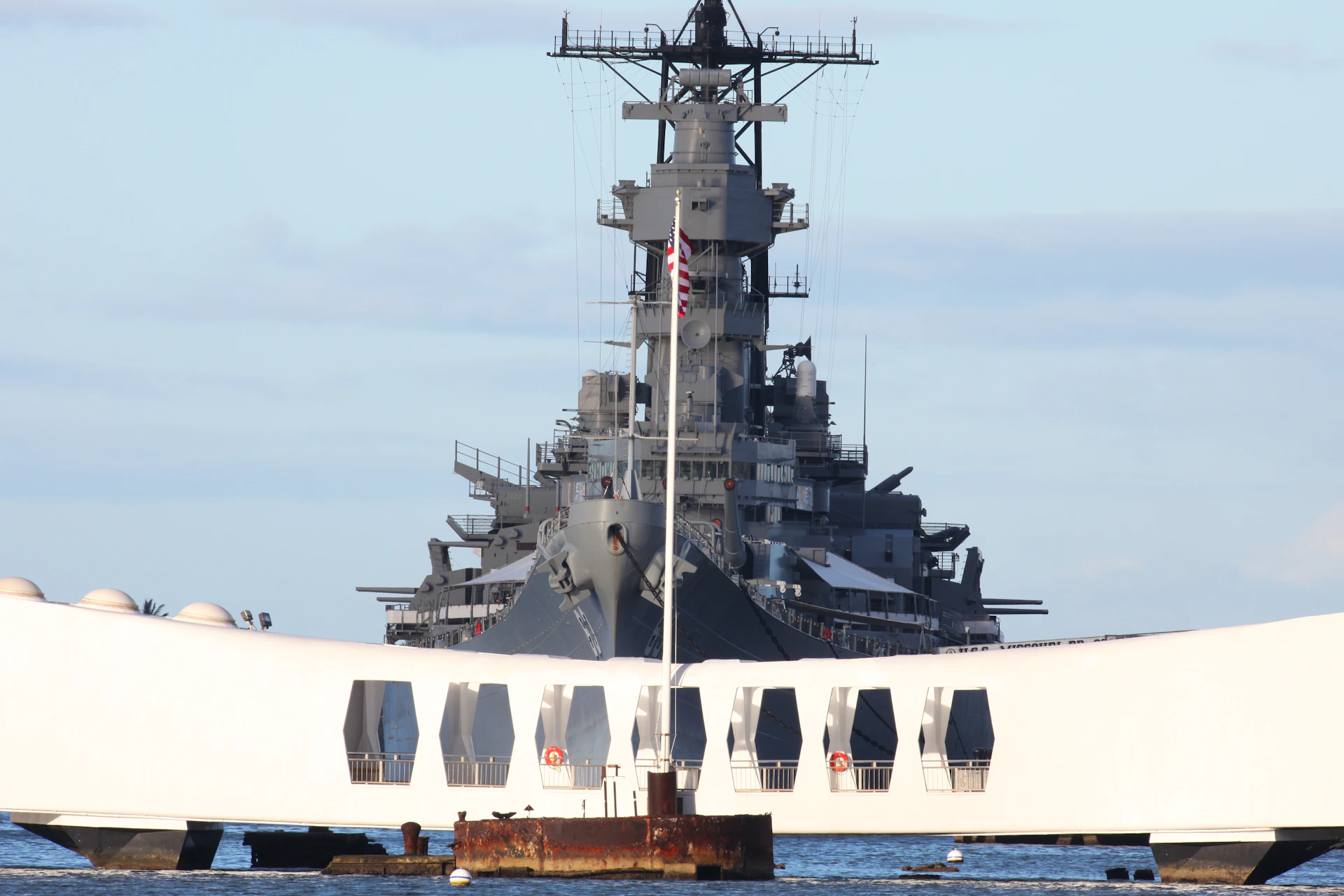 A large battleship is docked behind a white memorial structure with a visible American flag in the center.