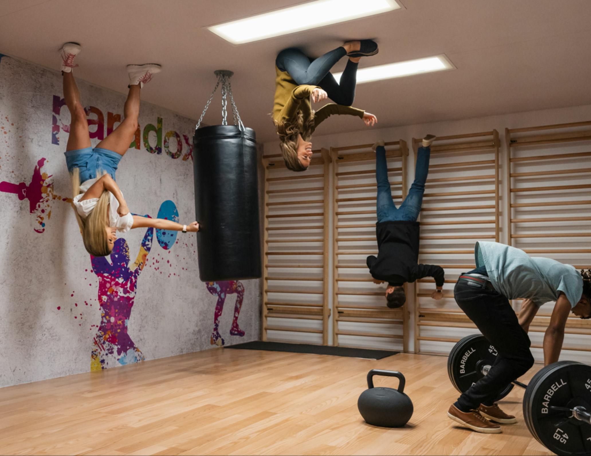 Des personnes semblent défier la gravité, se tenant au plafond dans une salle de sport avec un sac de frappe et des appareils de fitness au sol.