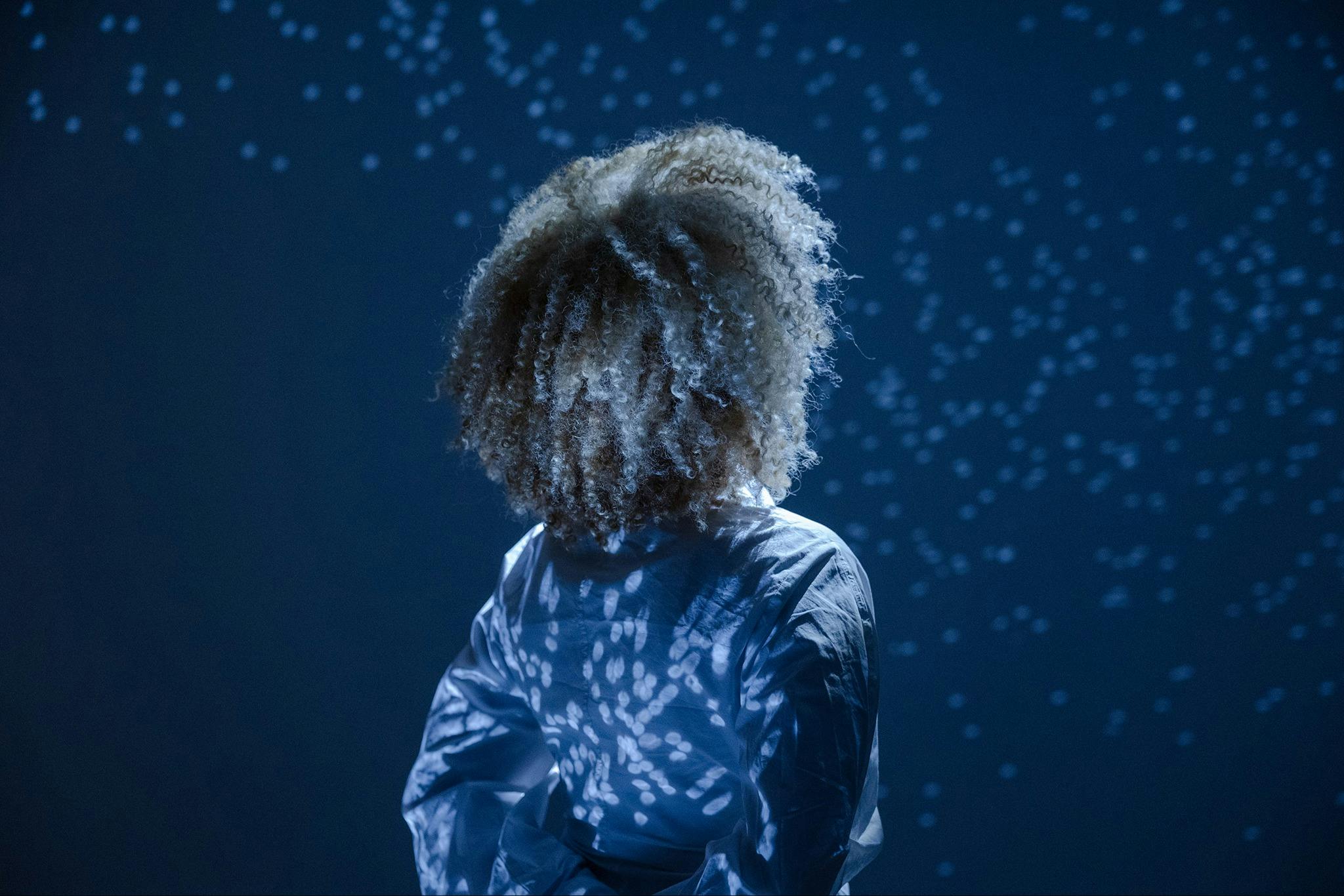 Persona con capelli ricci e camicia bianca in piedi davanti a uno sfondo blu con punti luce sparsi.