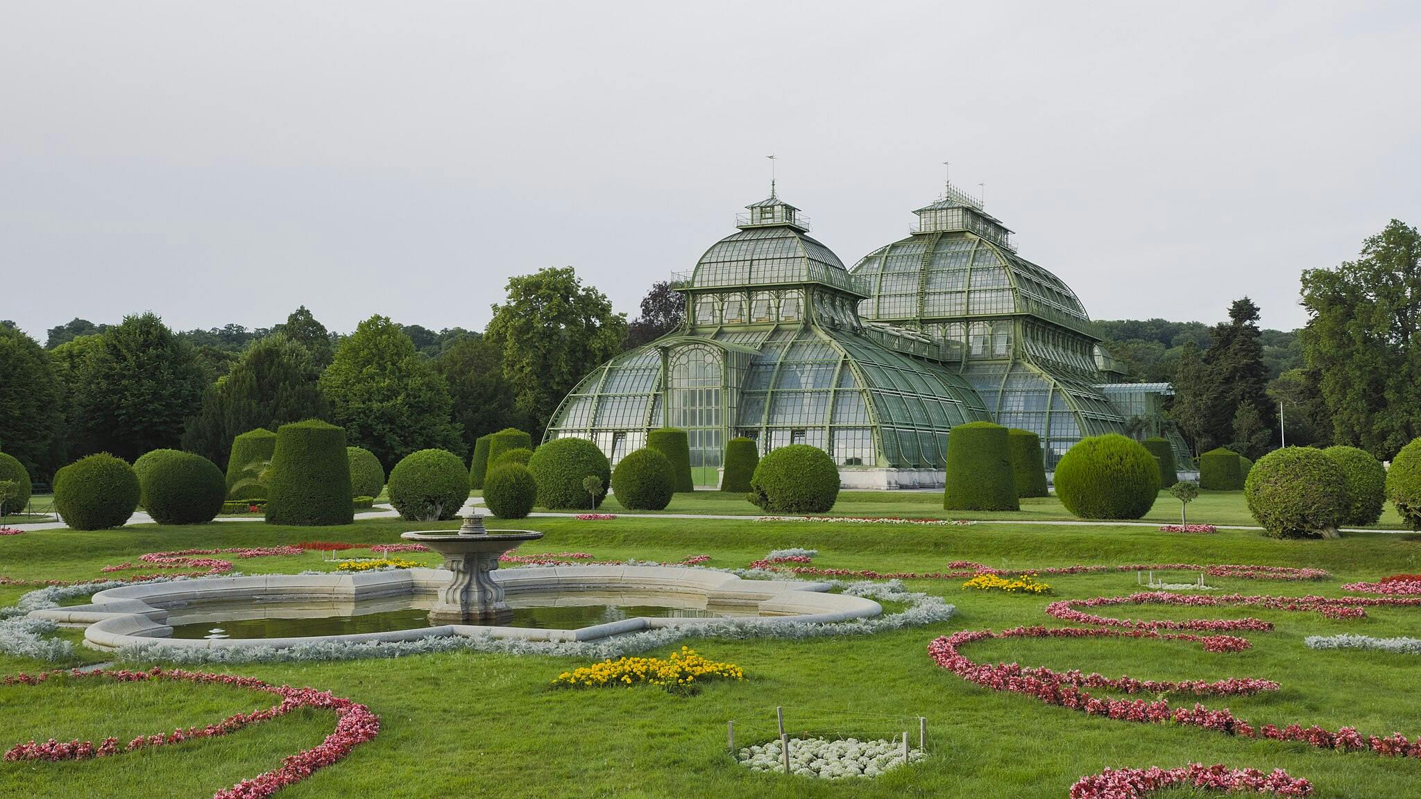 Duża szklana szklarnia z kopulastym dachem stoi w zadbanym ogrodzie z krzakami topiary, kolorowymi kwietnikami i fontanną.
