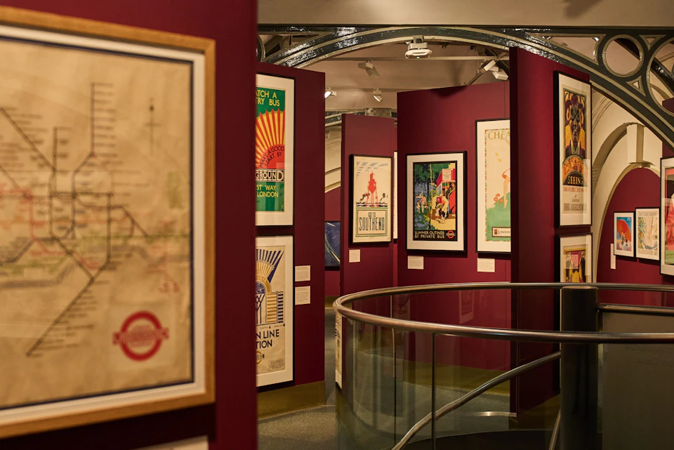 A museum exhibit featuring posters with vintage designs on deep red walls and a curved railing in the foreground.