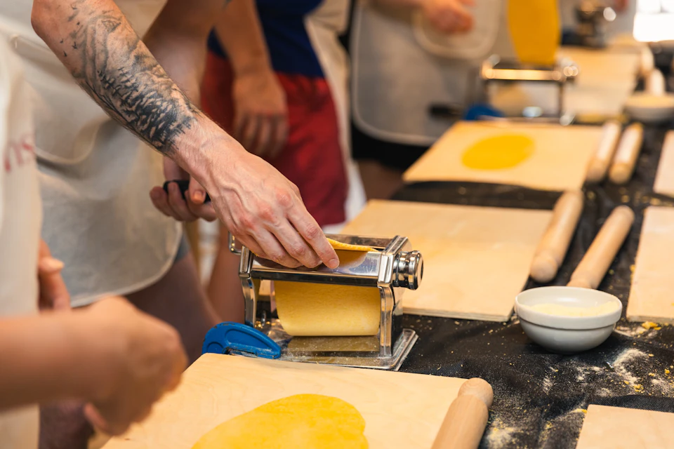 People using a pasta machine to roll out dough in a cooking class setting, with wooden boards and rolling pins visible.
