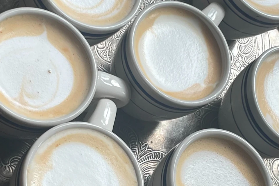 Several cups of cappuccino with frothy milk tops arranged closely together on a patterned metal tray.
