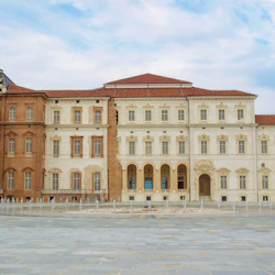 Utsmyckad byggnad med en blandning av rött och vitt tegel, välvda fönster, rött tegeltak och en fontän på en stor stenlagd innergård.