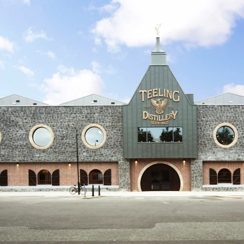 A modern distillery building with circular windows and an arched entrance. The sign reads "Teeling Distillery Newmarket."
