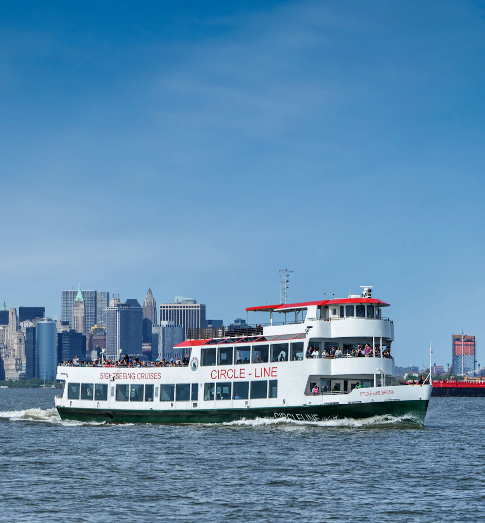 A sightseeing cruise boat labeled "CIRCLE LINE" sails on the water with a city skyline in the background under a clear blue sky.