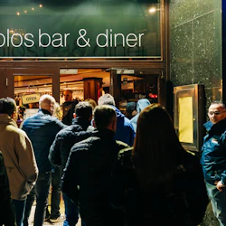 En folkmassa väntar utanför en bar och middag på kvällen, med en "öppen" skylt synlig på dörren.