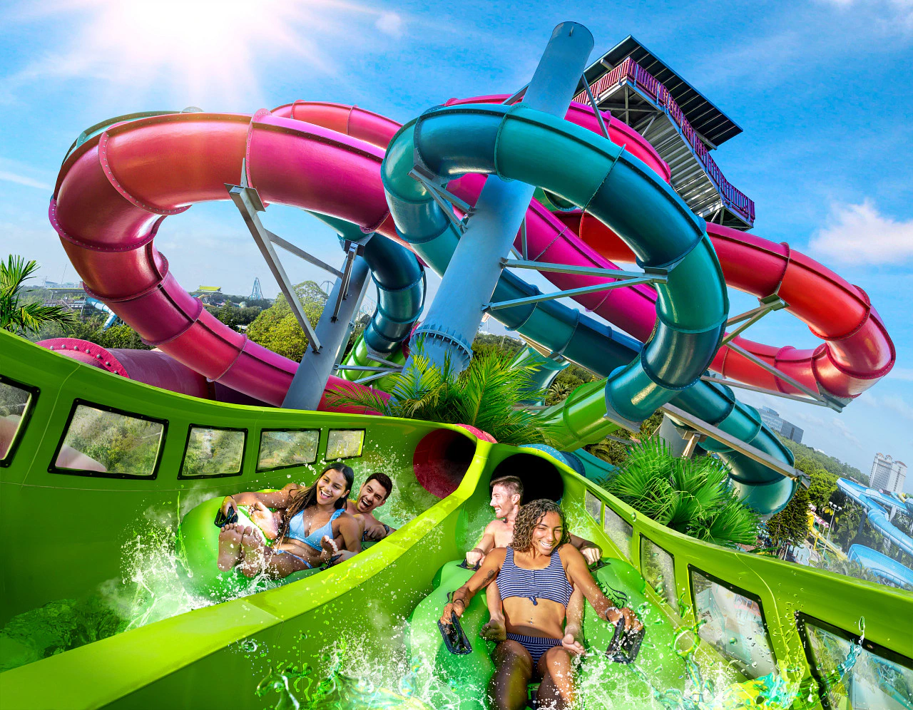 Four people descending a green waterslide with intertwined red, blue, and purple slides in the background under a clear blue sky.