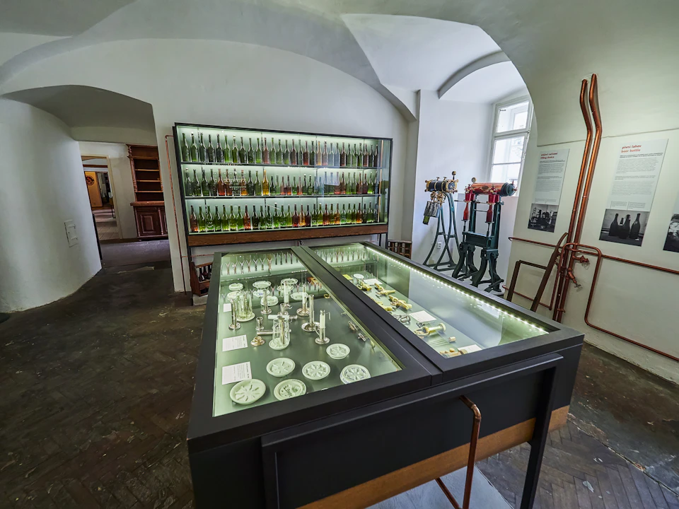 Museum exhibit room featuring glass cases with historical tools, machinery, and shelves of colorful bottles against white walls.
