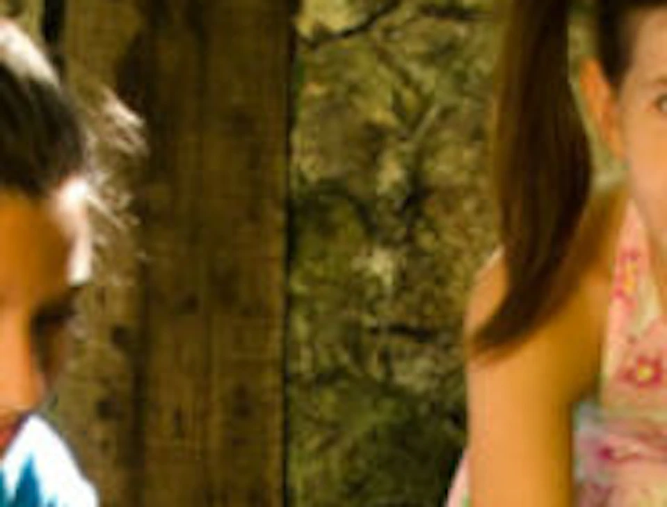 Two girls, one kneeling and one standing, are stroking a brown dog in an area with stone walls and a gravel floor.