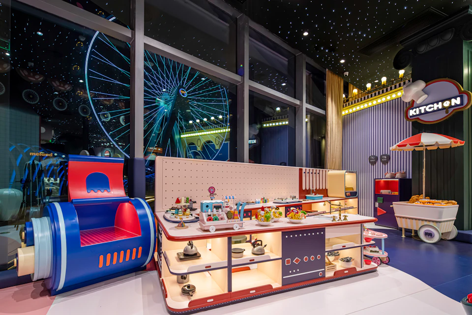 A colorful indoor play area with toy displays and a backdrop of a large ferris wheel visible through glass windows.