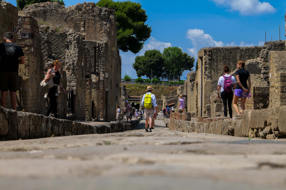 Tourists walking among ancient ruins on a stone pathway, with trees and a blue sky in the background.
