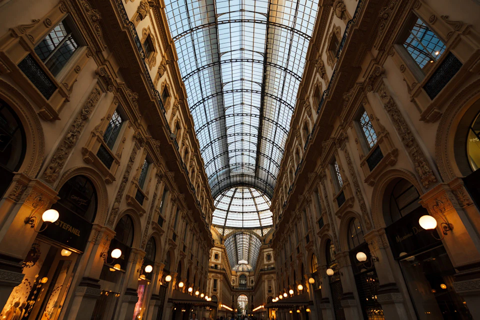 An elegant shopping arcade with a high arched glass ceiling, ornate architecture, and hanging lights along the walkway.