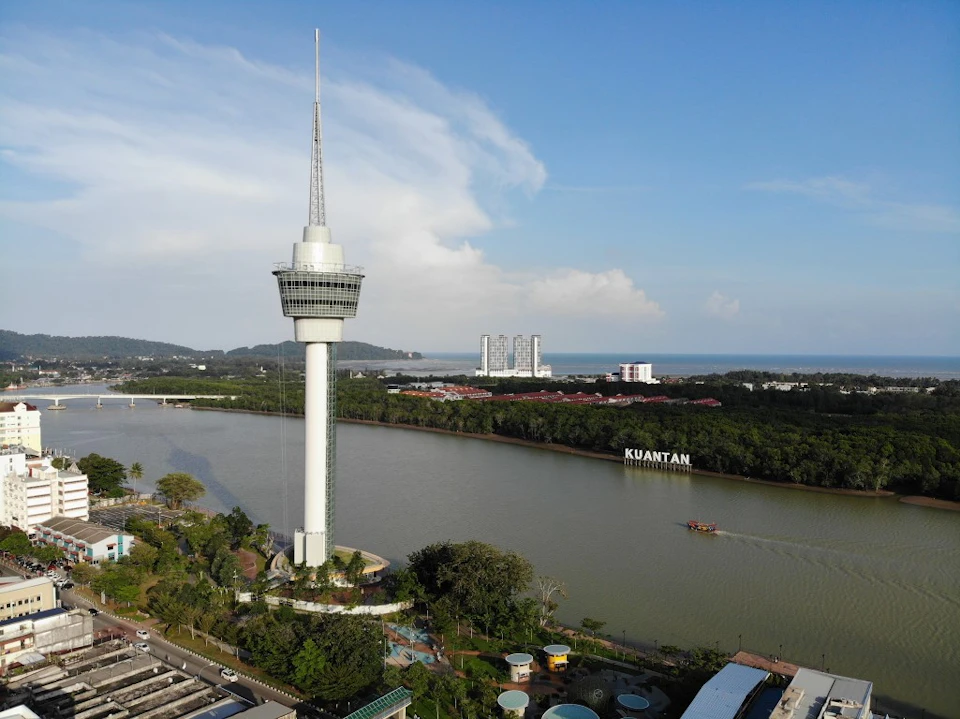 A tall observation tower by a river, with the word 'Kuantan' visible across the water, surrounded by greenery and buildings.
