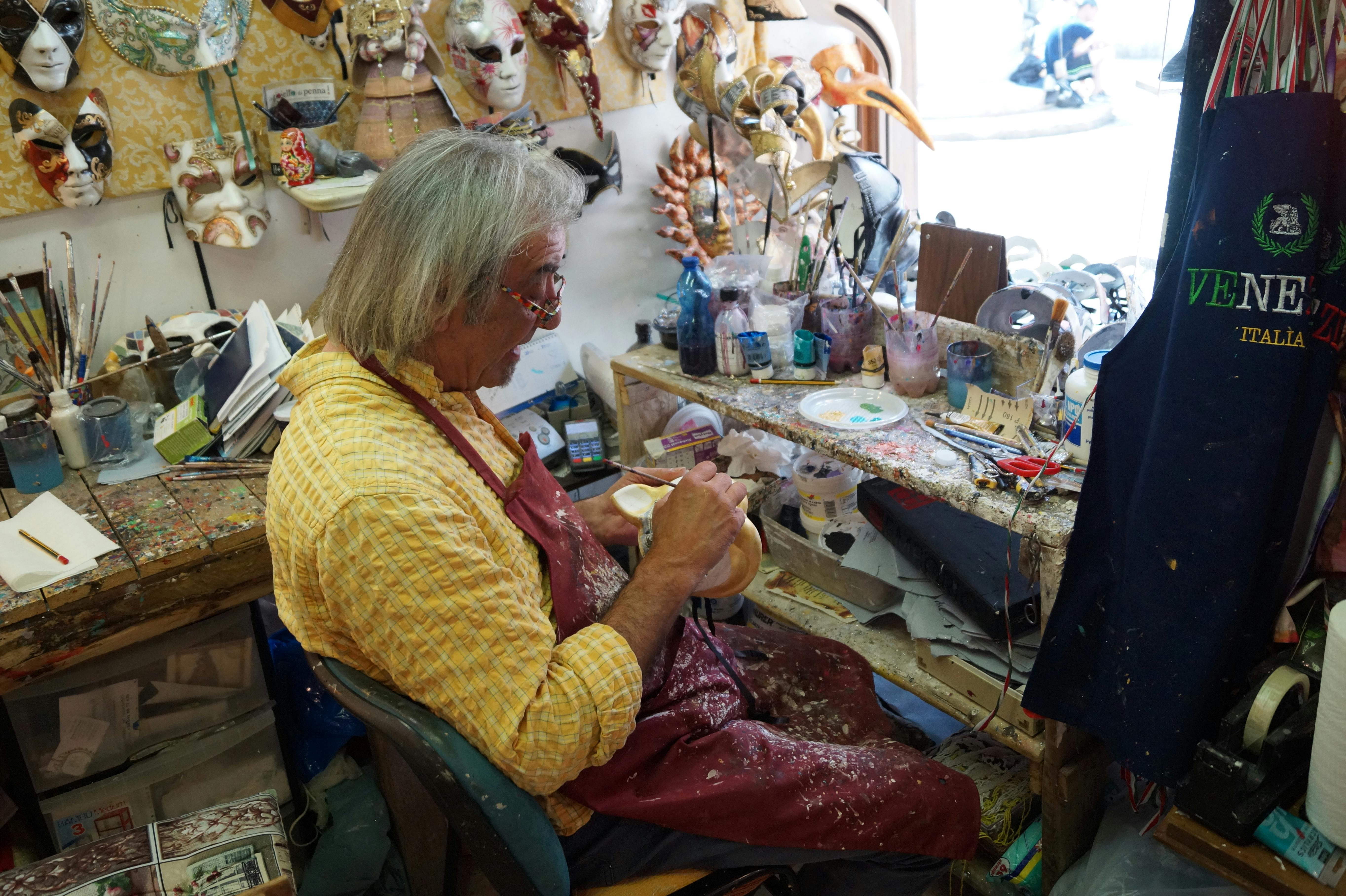 노란색 셔츠와 빨간색 앞치마를 입은 사람이 공구, 페인트, 마스크로 가득 찬 공방에서 마스크를 제작합니다.