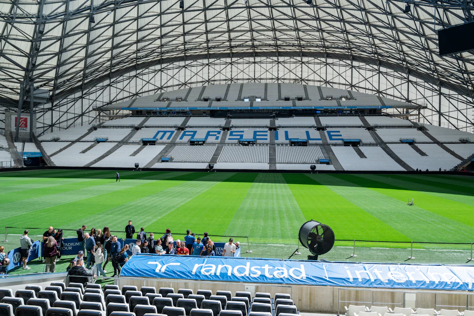 A large football stadium with "MARSEILLE" written on the seats, a green field, and people gathered near the foreground.