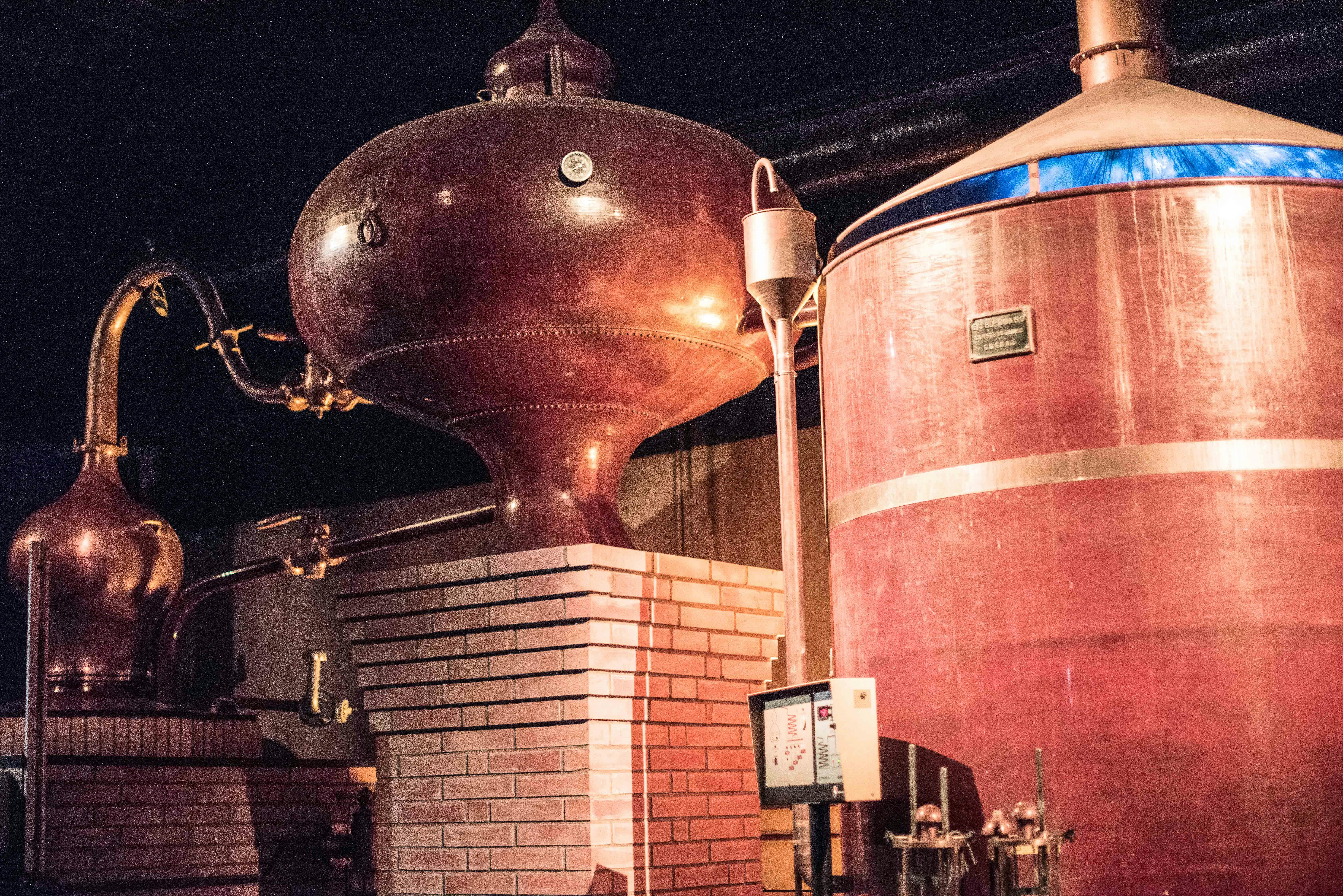 Grans dipòsits de destil·lació de coure amb bases de maó a l'interior d'una destil·leria, amb canonades i un quadre de control.