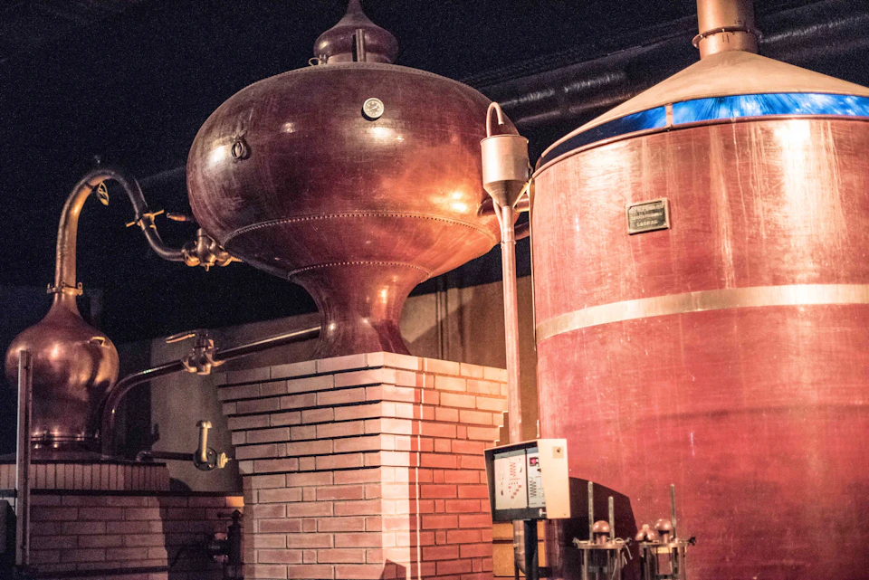 Large copper distillation tanks with brick bases inside a distillery, featuring pipes and a control panel.