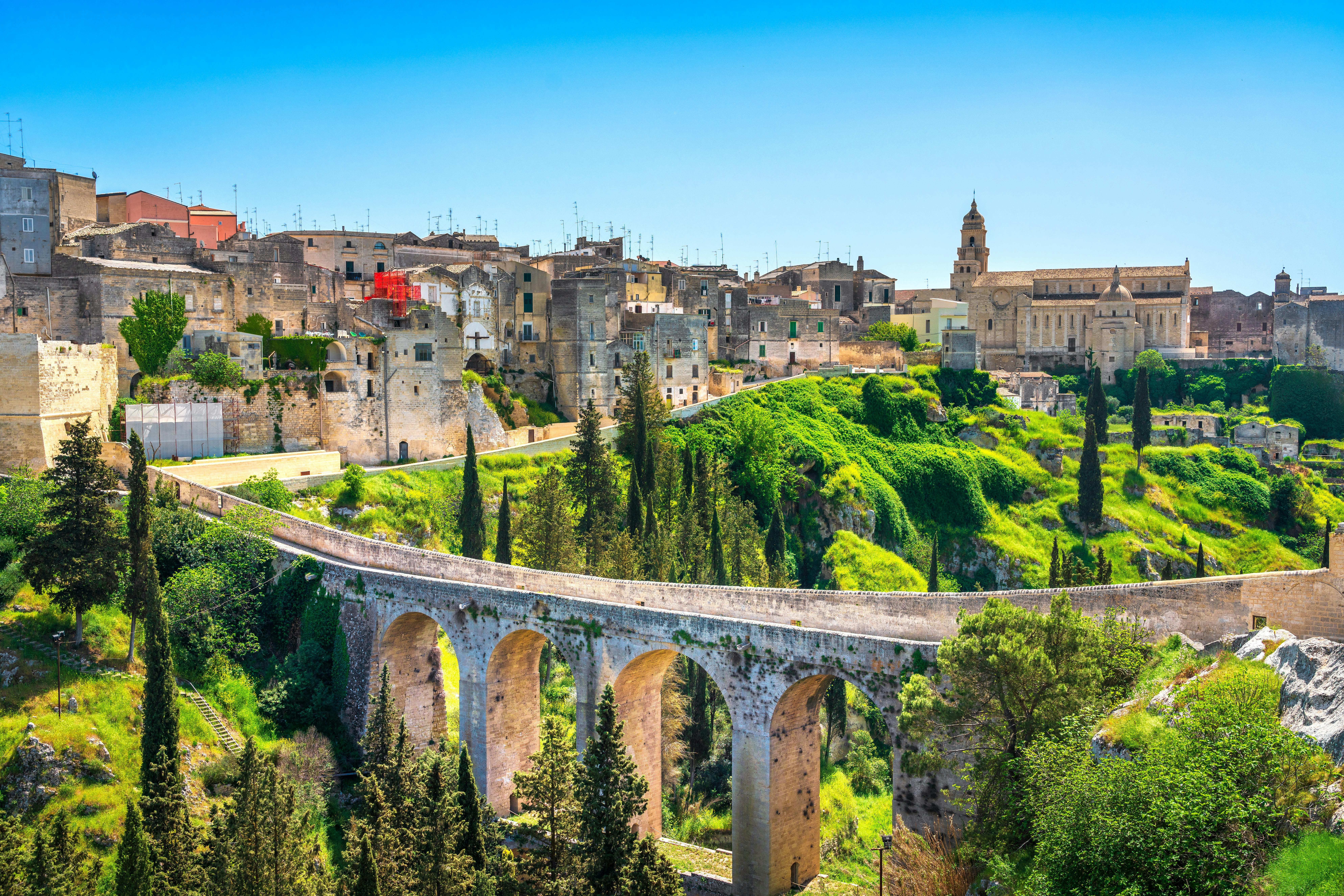 A historic stone bridge spans a green valley, leading to a town with ancient buildings and a bell tower under a clear blue sky.