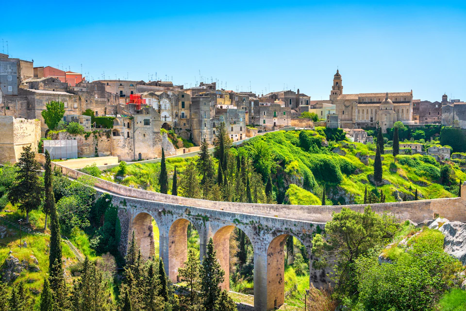 A historic stone bridge spans a green valley, leading to a town with ancient buildings and a bell tower under a clear blue sky.