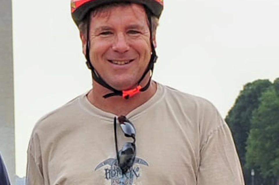 A group of people wearing helmets stand beside their bicycles on a grass field with the Washington Monument in the background.