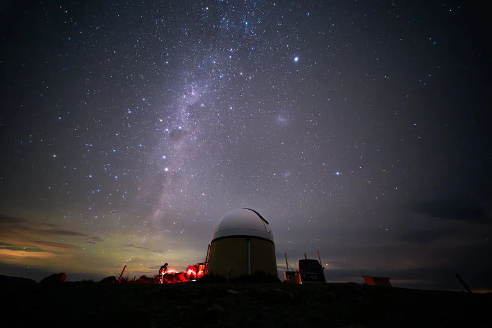 A night sky filled with stars and the Milky Way above an observatory dome and people with red lights on a hill.