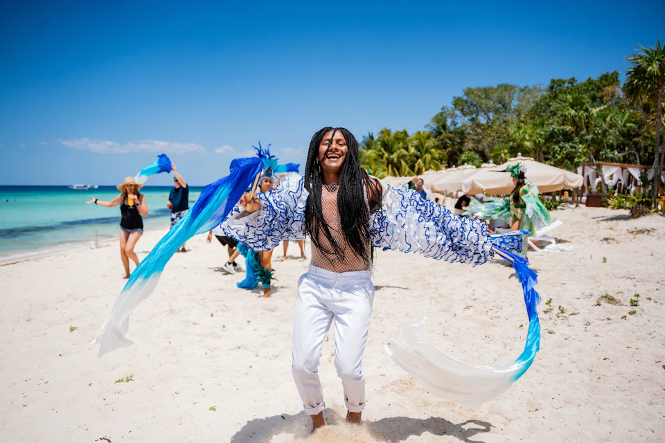 A person joyfully dances on a beach in white pants and a decorative top, surrounded by others with blue and green accessories.