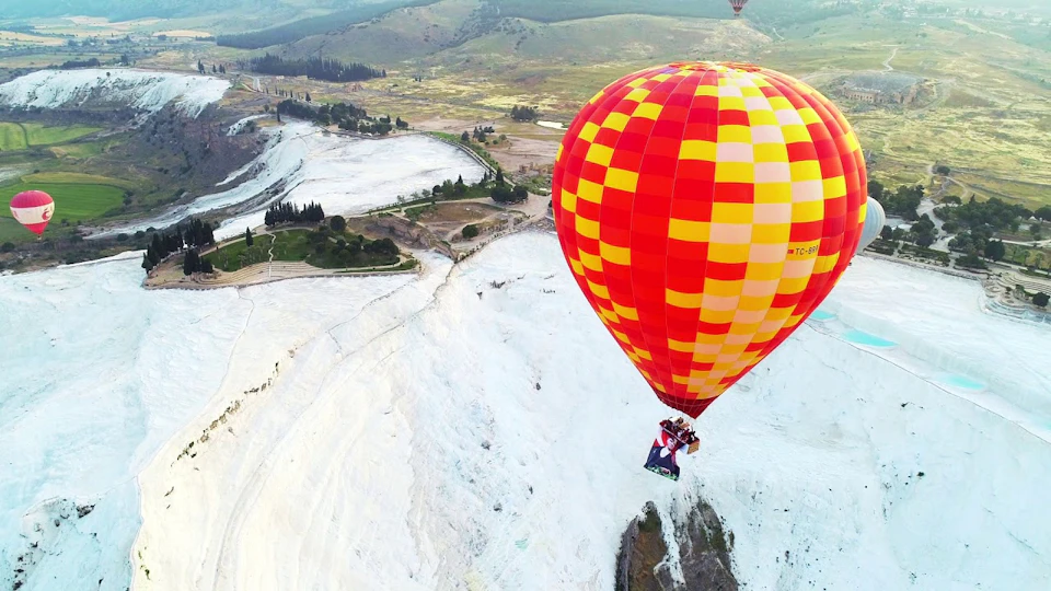 A red and yellow hot air balloon with two passengers flying over a landscape with white formations and green fields.