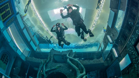 Divers in an indoor pool with underwater structures, including a tree-like sculpture and windowed buildings.