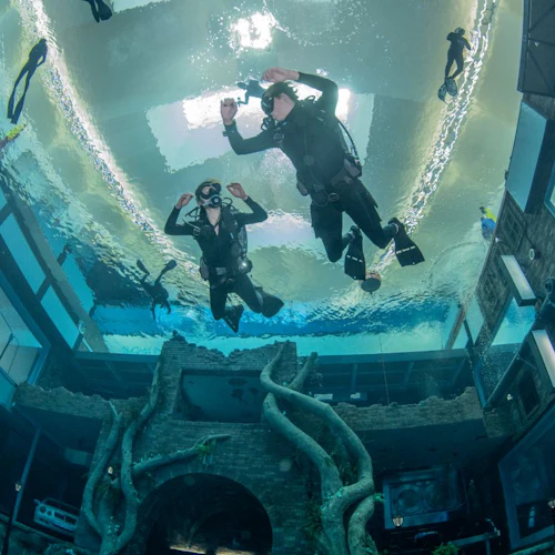Divers in an indoor pool with underwater structures, including a tree-like sculpture and windowed buildings.