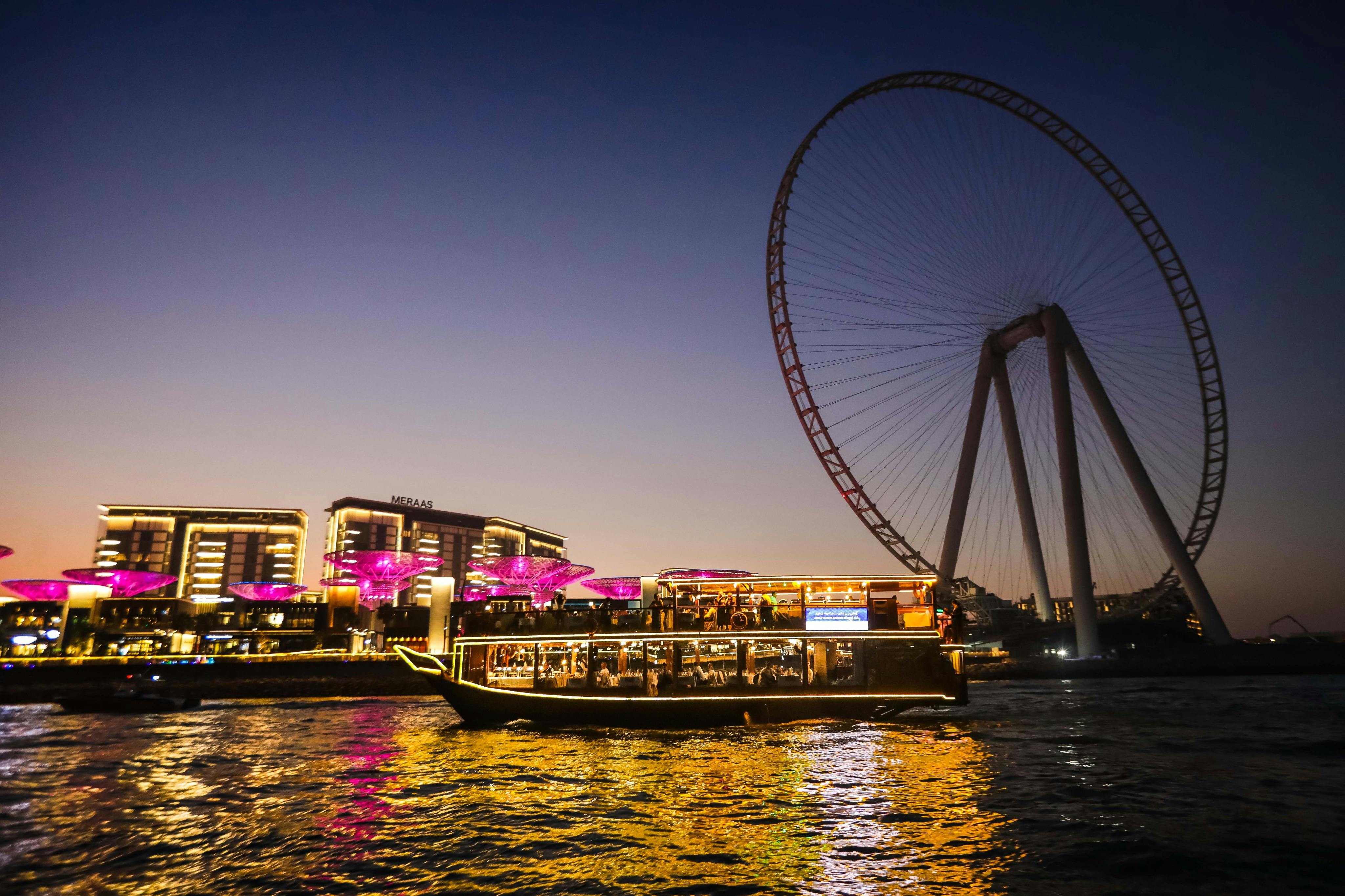 Beleuchtetes Boot auf dem Wasser in der Abenddämmerung, großes Riesenrad, Gebäude und rosa beleuchtete Strukturen im Hintergrund.