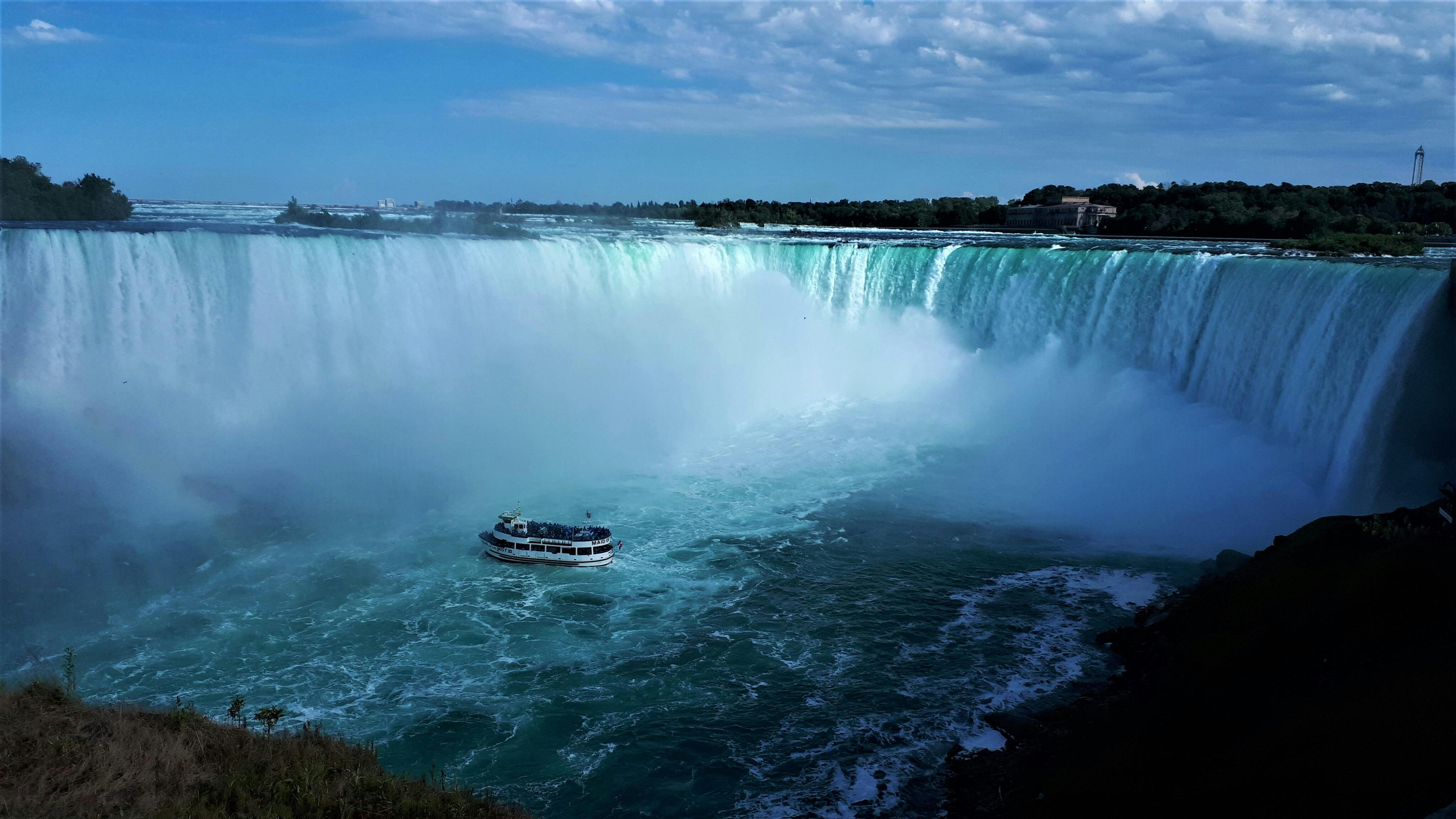 A sightseeing boat approaches the base of a large, misty waterfall under a partly cloudy sky.