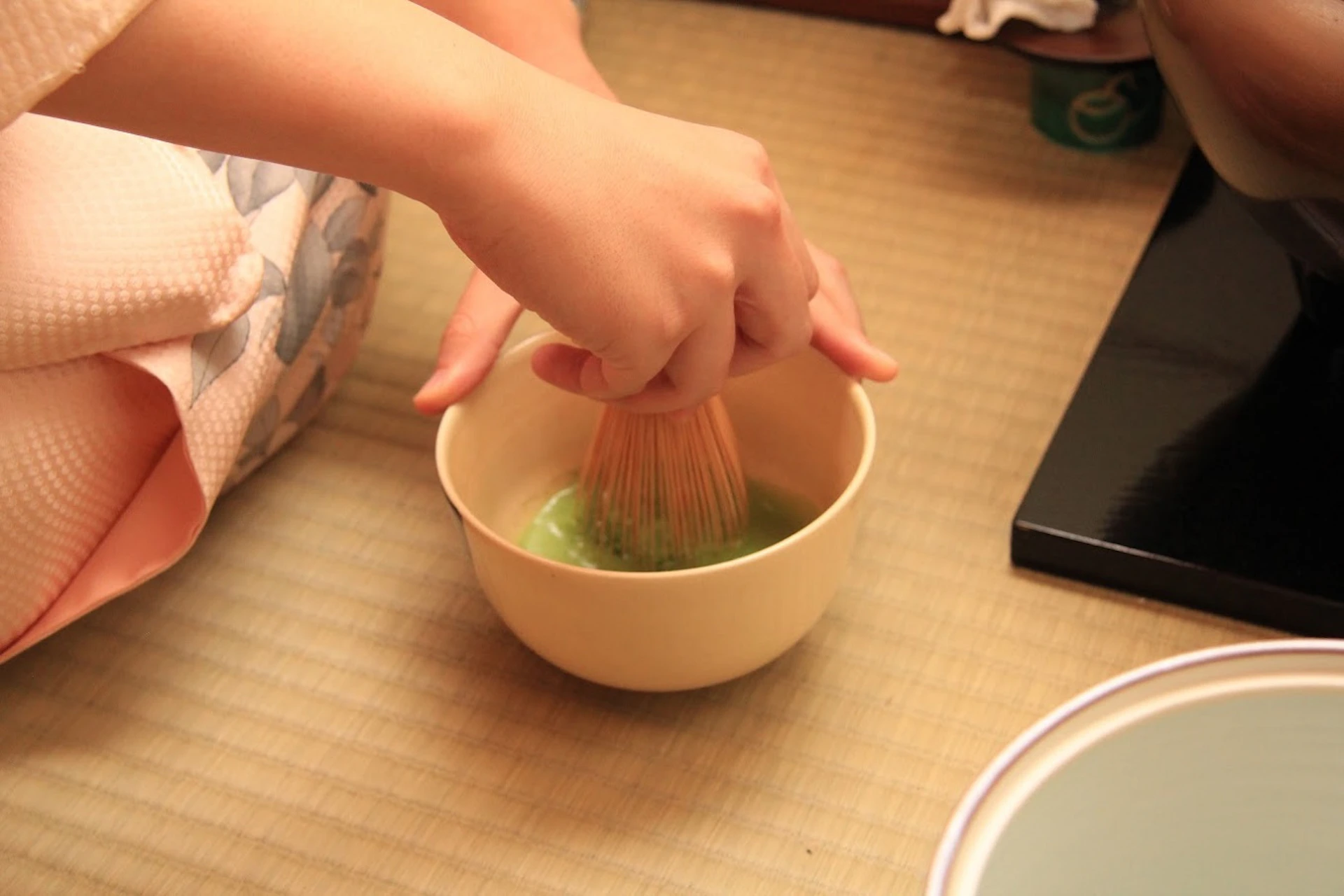 Hands whisking matcha in a bowl on a tatami mat.