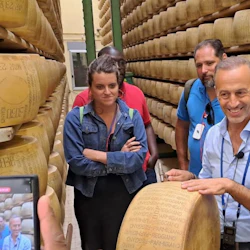 A group of people stands in an aisle between shelves stacked with large wheels of cheese, one man smiling and holding a cheese wheel.