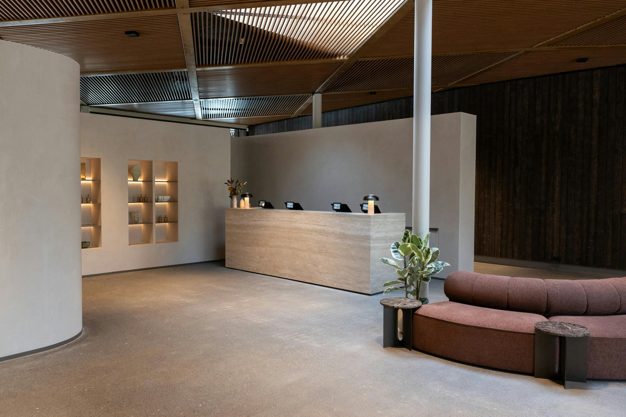 Modern reception area with a long, light-colored desk, several mounted tablets, a brown couch, potted plants, and wooden ceiling panels.