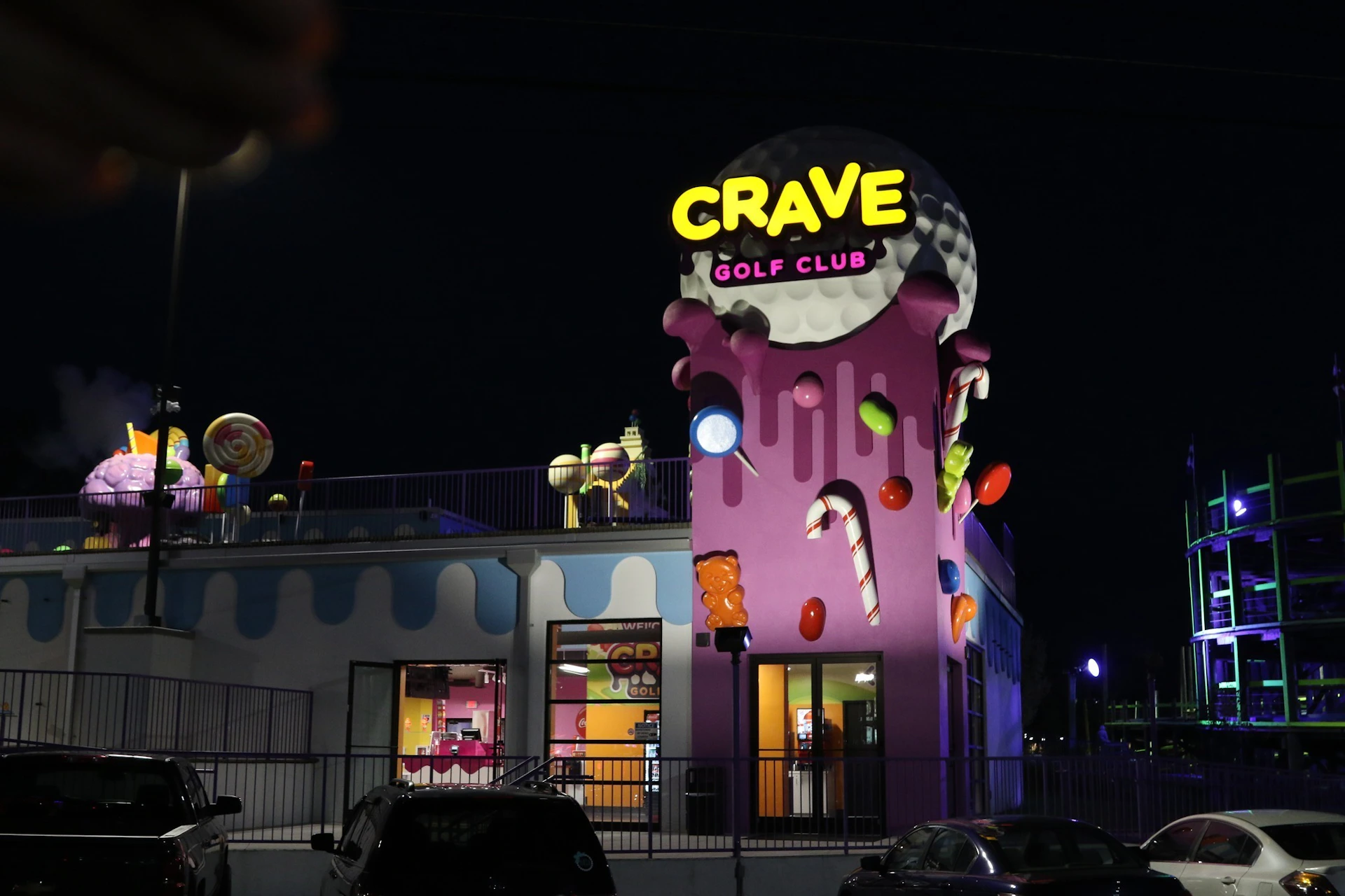 A brightly lit building at night with "CRAVE GOLF CLUB" signage, decorated with candy shapes and an ice cream theme.