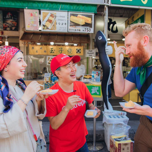 Three people are smiling and eating food with chopsticks outside a market stall with colorful signs and displays.