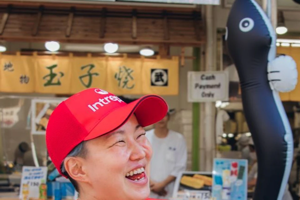 Three people are smiling and eating food with chopsticks outside a market stall with colorful signs and displays.