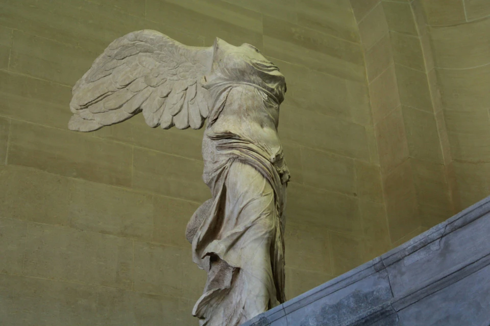 A marble statue of a winged, headless figure, draped in flowing garments, displayed in a museum setting.