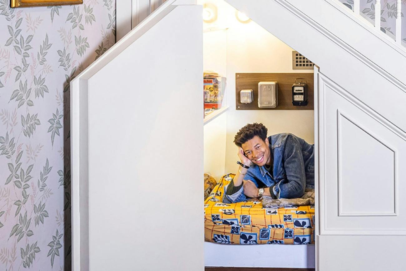 A person smiles while lying on a bed in a cozy space under stairs, decorated with a yellow patterned blanket and shelves.