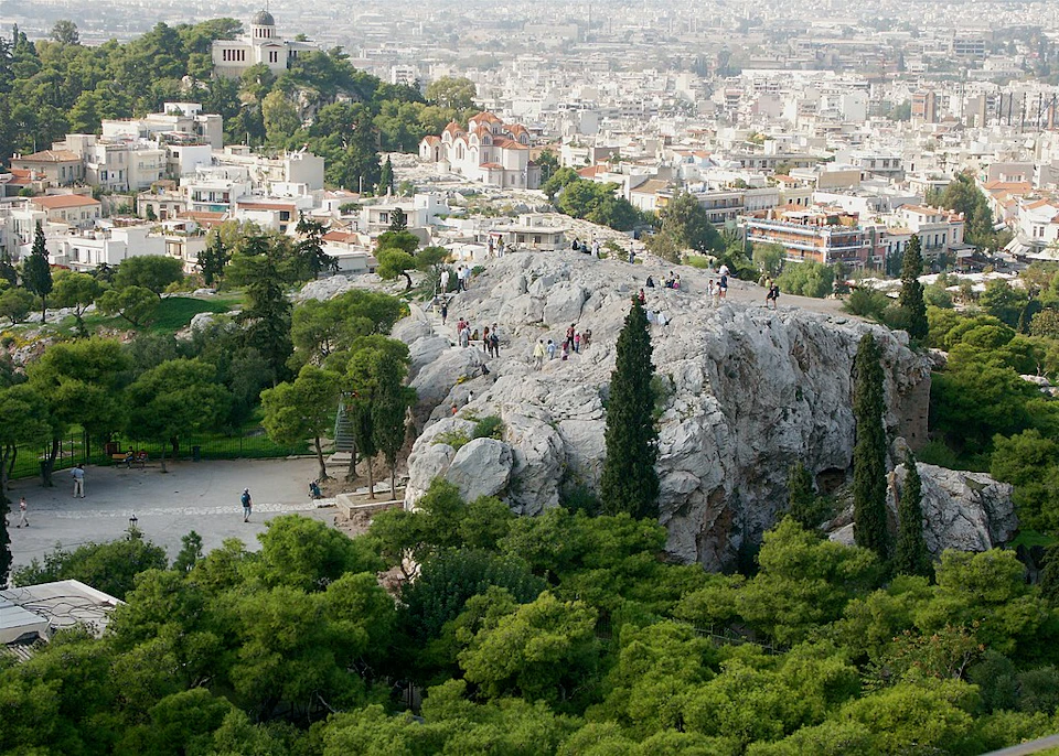 People walking on a large rock formation with a cityscape and greenery in the background.