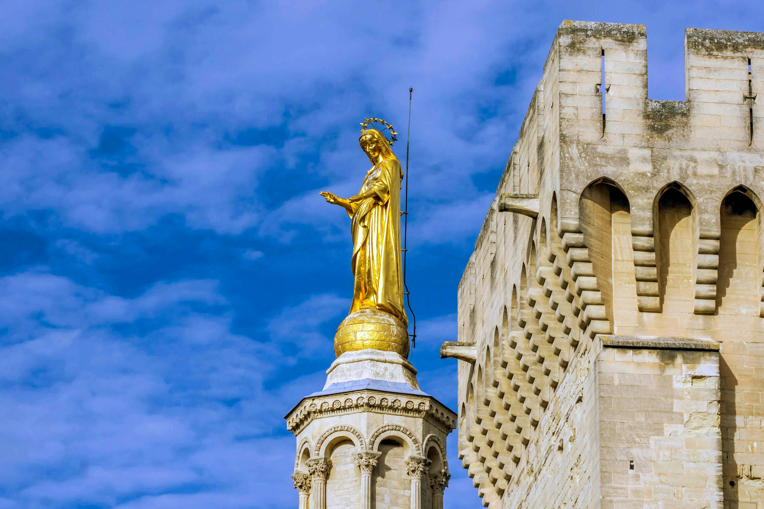 A golden statue with a halo stands atop an ornate column beside a stone building under a blue sky with scattered clouds.