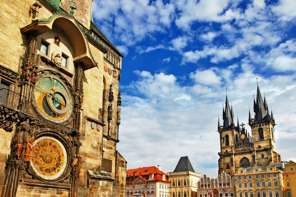 A historic astronomical clock on a building next to gothic-style towers and ornate structures under a bright blue sky with white clouds.