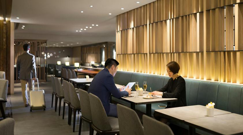 Two people seated in a modern restaurant with wooden panel walls, teal seating, and tables with glasses and small vases.