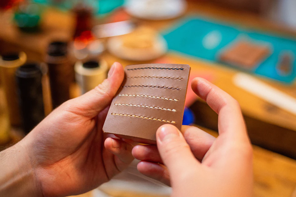 Hands holding a leather sample with different stitching patterns in a workshop setting.