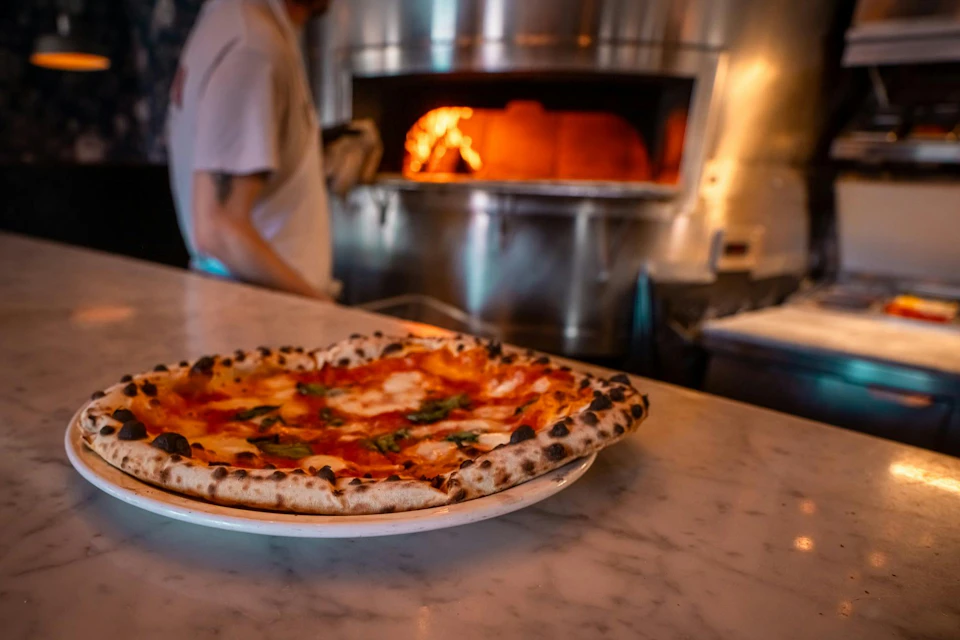 A freshly baked pizza on a marble counter with a wood-fired oven in the background and a person partially visible.