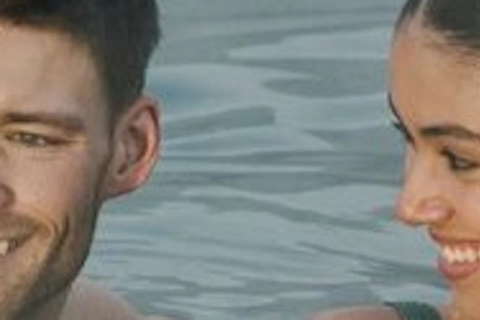 Two people relaxing in a steaming outdoor pool near a modern, arched building in a snowy landscape.