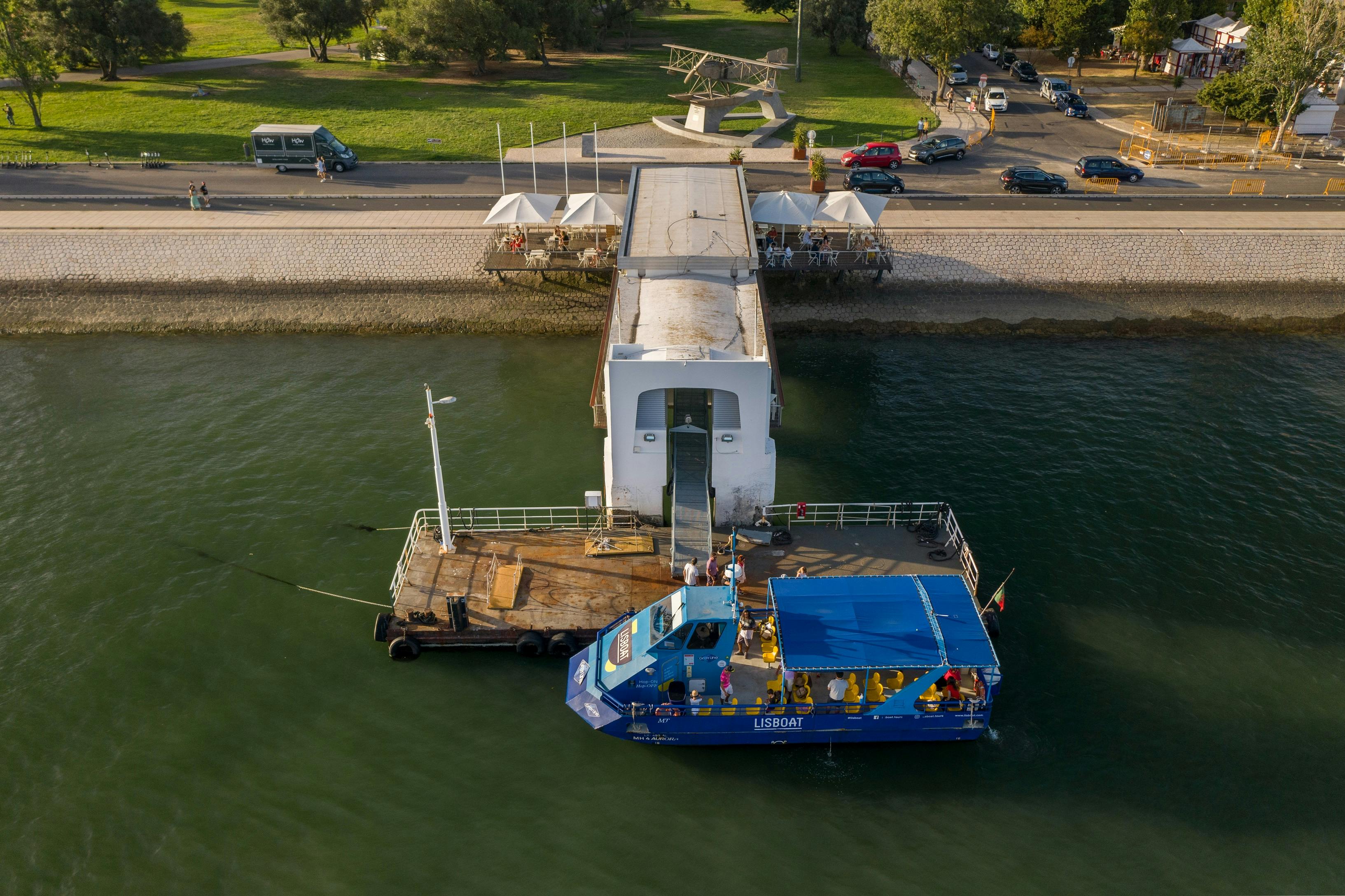 사람들이 탑승하는 부두에 배가 정박했습니다. 부두는 주차된 차량과 인근 나무가 있는 강변 공원에서 뻗어 있습니다.