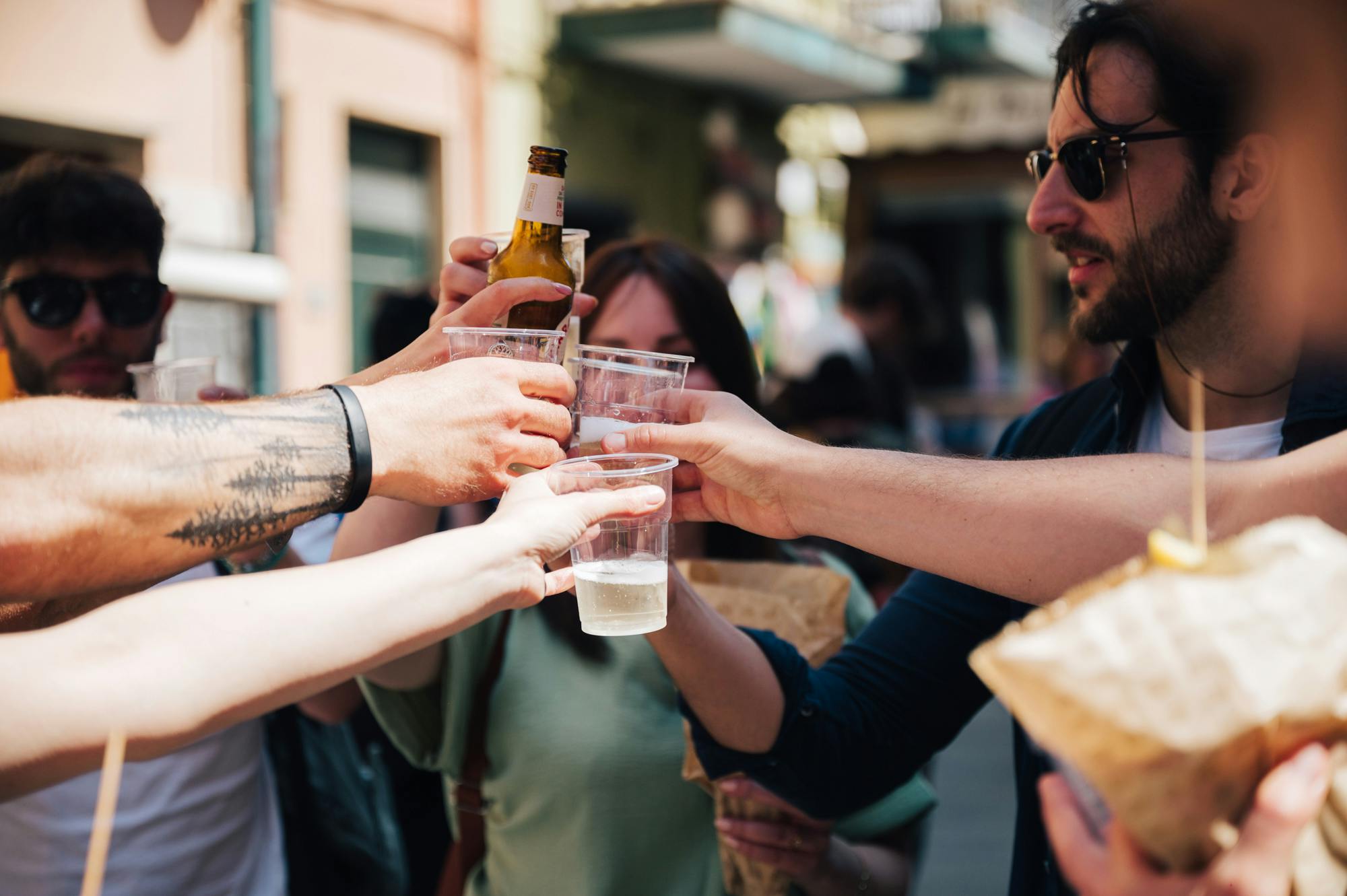 人们在户外环境下碰撞塑料杯和塑料瓶。 一个人拿着一袋食物。 人脸部分可见。