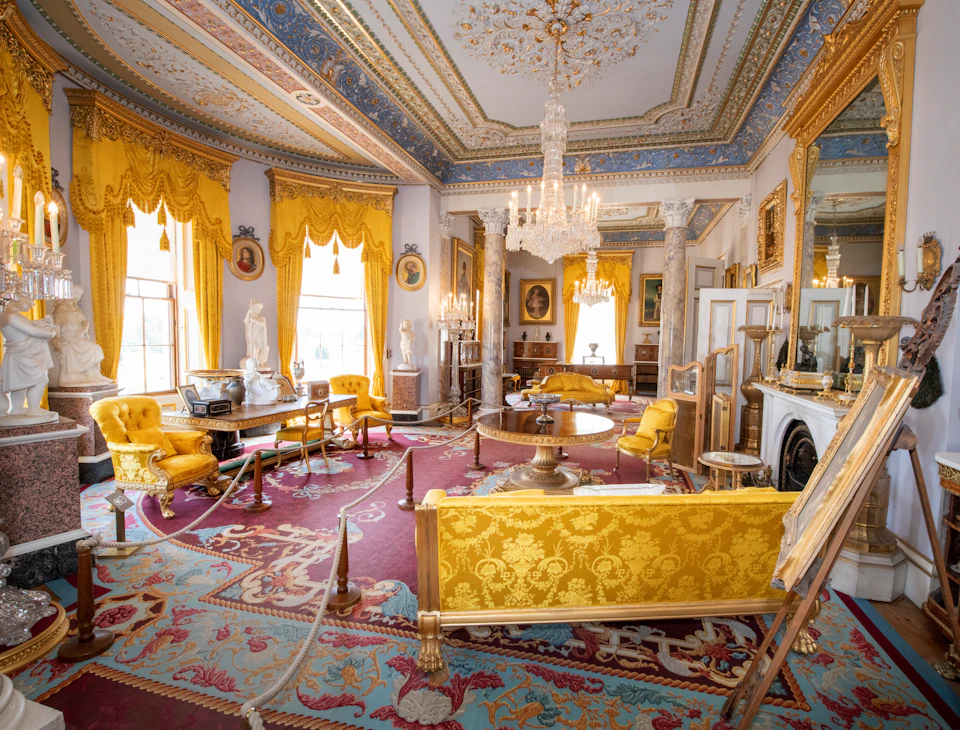 Luxurious room with ornate décor, yellow drapes and furniture, chandeliers, statues, framed paintings, and antique furnishings.